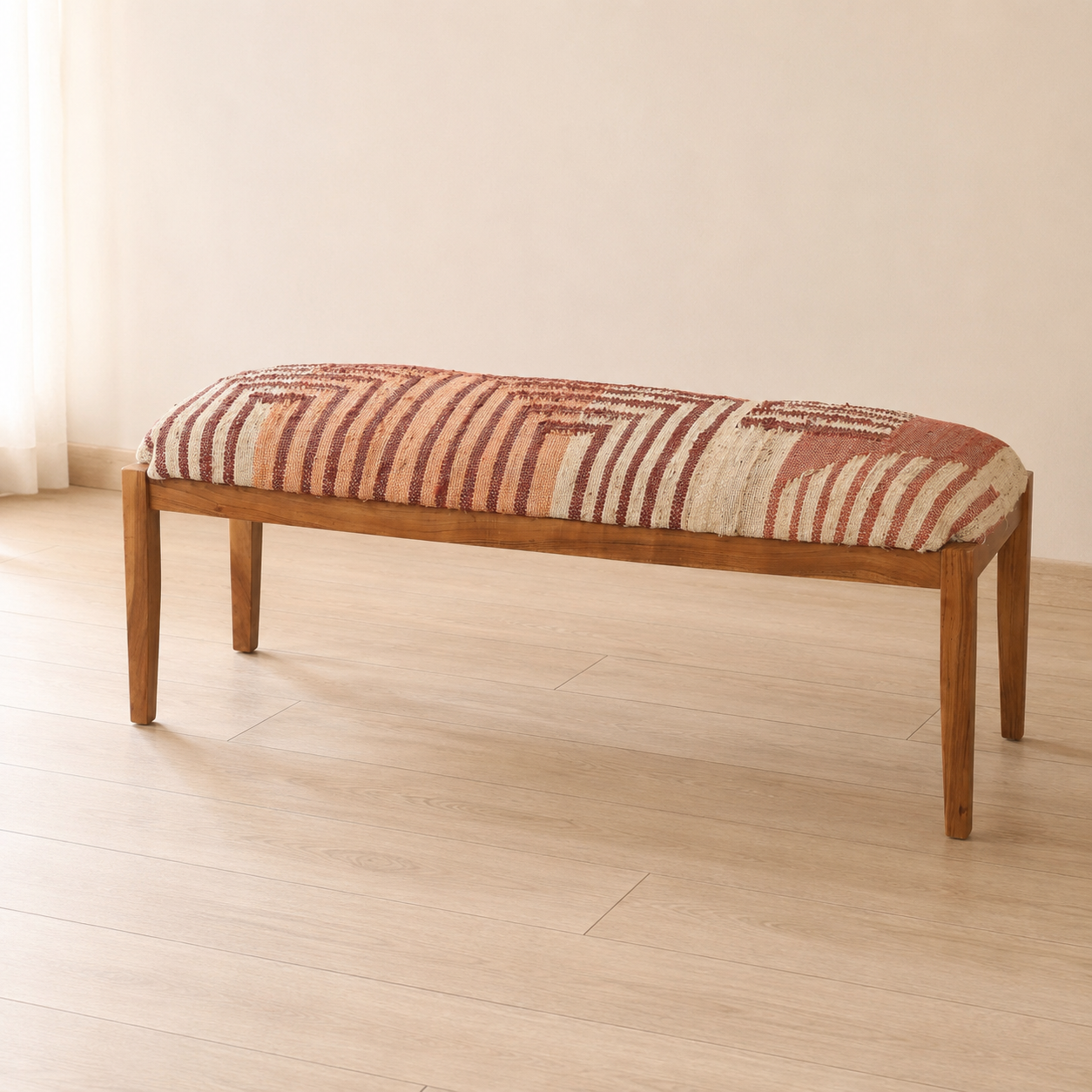 A wooden kilim ottoman bench with a rectangular cushion upholstered in a rust red, beige, and cream striped patterned fabric, set on a light wood floor against a neutral cream wall.