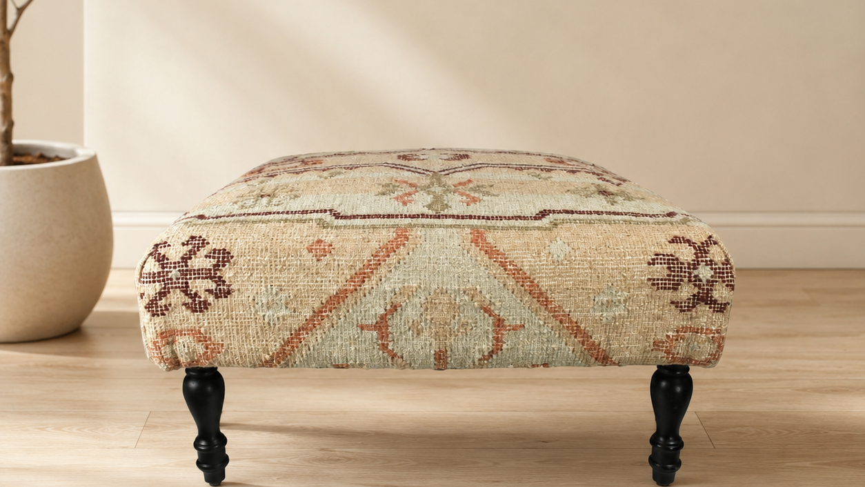 A square upholstered ottoman stool with a beige and rust patterned rug-style fabric top and four dark turned legs, shown in a softly lit room with a wood floor and potted plant.