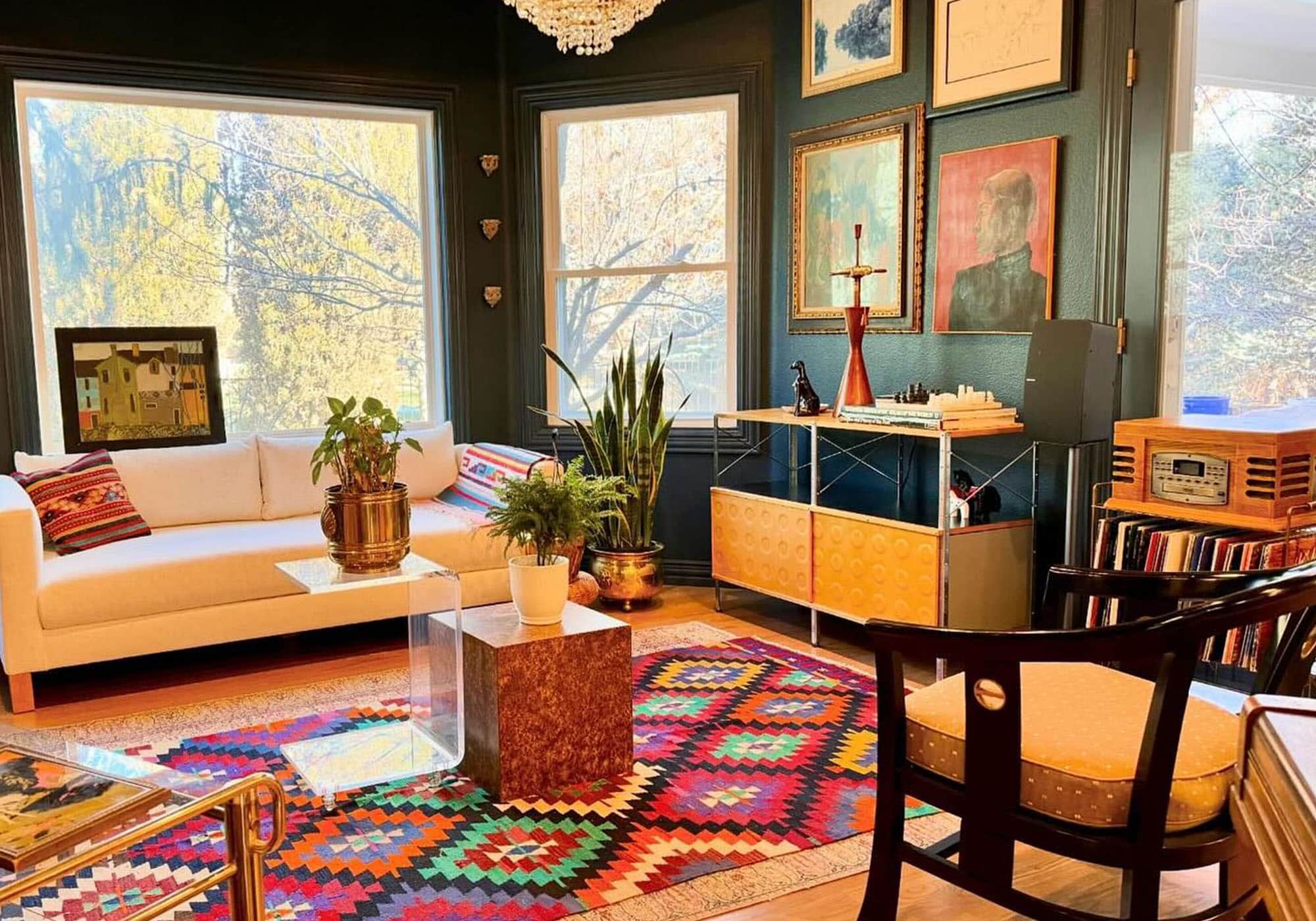 A cozy, eclectic living room featuring a vibrant antique kilim rug purchased from Kilim Couture. The rug’s bold geometric patterns in red, blue, and green complement the modern white sofa adorned with colorful woven pillows. Dark green walls showcase an artful gallery, while mid-century furniture, lush plants, and warm lighting create an inviting atmosphere. A beautiful showcase of a Kilim Couture rug in a happy customer's home.