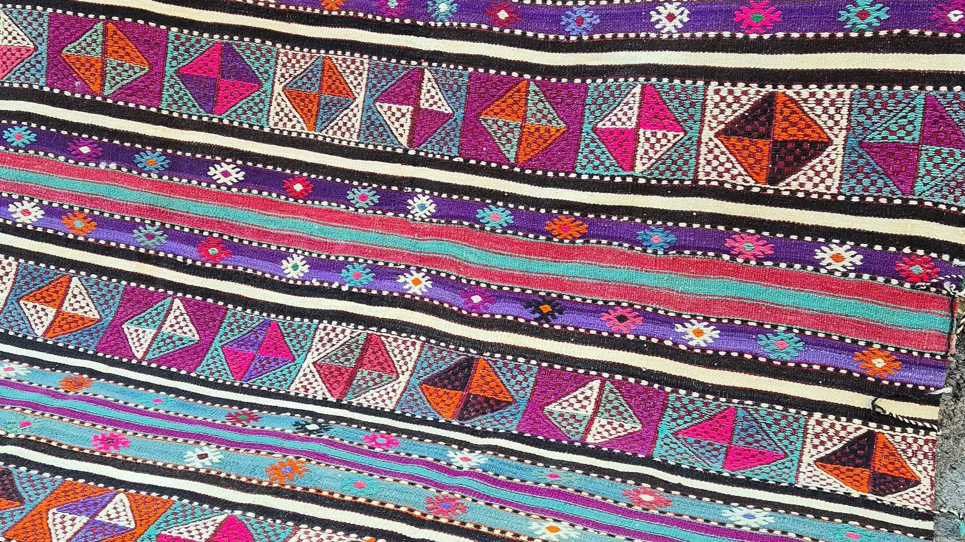 Vintage Turkish Balikesir Striped Kilim. Handwoven in the 1960s in Balikesir, this 5x8 semi-antique kilim is a rare gem of Anatolian textile heritage. Featuring bold tribal stripes and symbolic 'burdock' and 'love and unison' motifs, its inky black and ivory base is accented by plum velvet, raspberry, turquoise, and desert rose. A sophisticated yet spirited heirloom, perfect for grounding interiors with artistic depth and bohemian elegance.