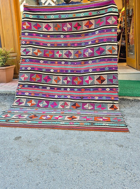 Vintage Turkish Balikesir Striped Kilim. Handwoven in the 1960s in Balikesir, this 5x8 semi-antique kilim is a rare gem of Anatolian textile heritage. Featuring bold tribal stripes and symbolic 'burdock' and 'love and unison' motifs, its inky black and ivory base is accented by plum velvet, raspberry, turquoise, and desert rose. A sophisticated yet spirited heirloom, perfect for grounding interiors with artistic depth and bohemian elegance.