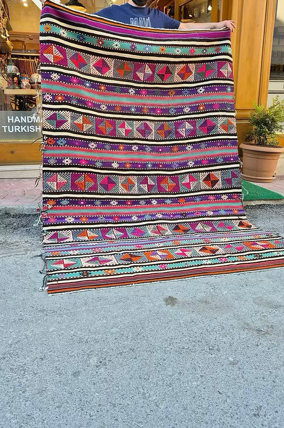 Vintage Turkish Balikesir Striped Kilim. Handwoven in the 1960s in Balikesir, this 5x8 semi-antique kilim is a rare gem of Anatolian textile heritage. Featuring bold tribal stripes and symbolic 'burdock' and 'love and unison' motifs, its inky black and ivory base is accented by plum velvet, raspberry, turquoise, and desert rose. A sophisticated yet spirited heirloom, perfect for grounding interiors with artistic depth and bohemian elegance.