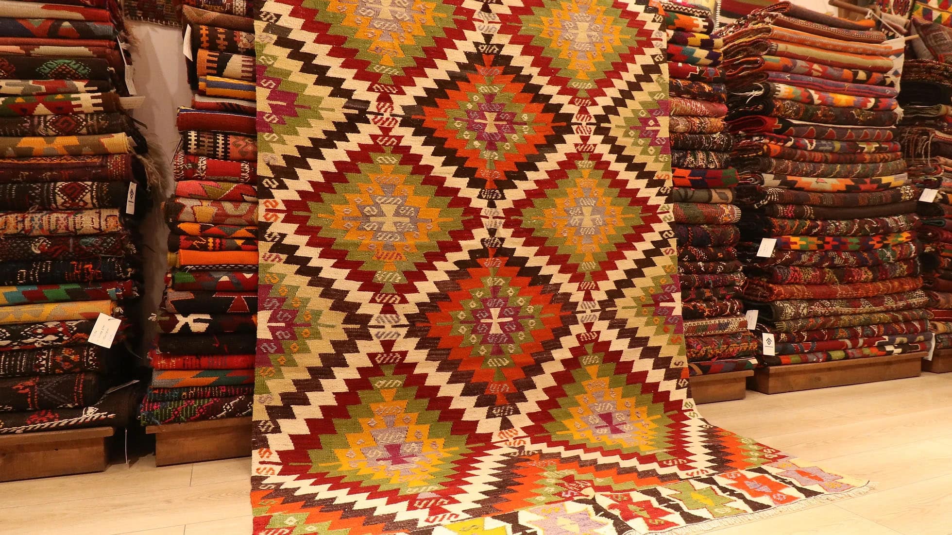 1960s handwoven kilim rug from Manisa, Turkey, 5x8 ft, featuring bold diamond motifs in pistachio, saffron, and clementine hues outlined in black on a white backdrop. The rug includes 'fetter' border motifs symbolizing connection and unity, with delicate tassels, blending cultural heritage with modern design.