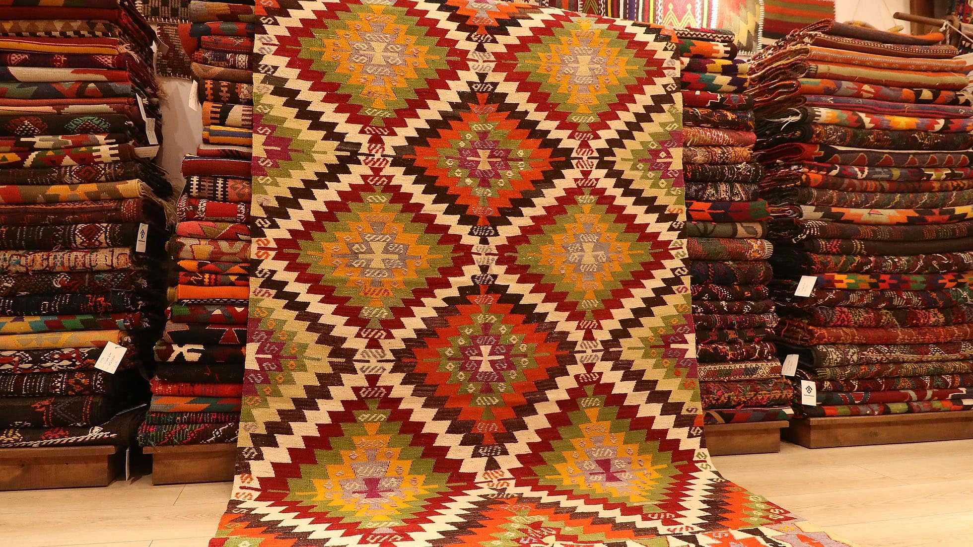 1960s handwoven kilim rug from Manisa, Turkey, 5x8 ft, featuring bold diamond motifs in pistachio, saffron, and clementine hues outlined in black on a white backdrop. The rug includes 'fetter' border motifs symbolizing connection and unity, with delicate tassels, blending cultural heritage with modern design.