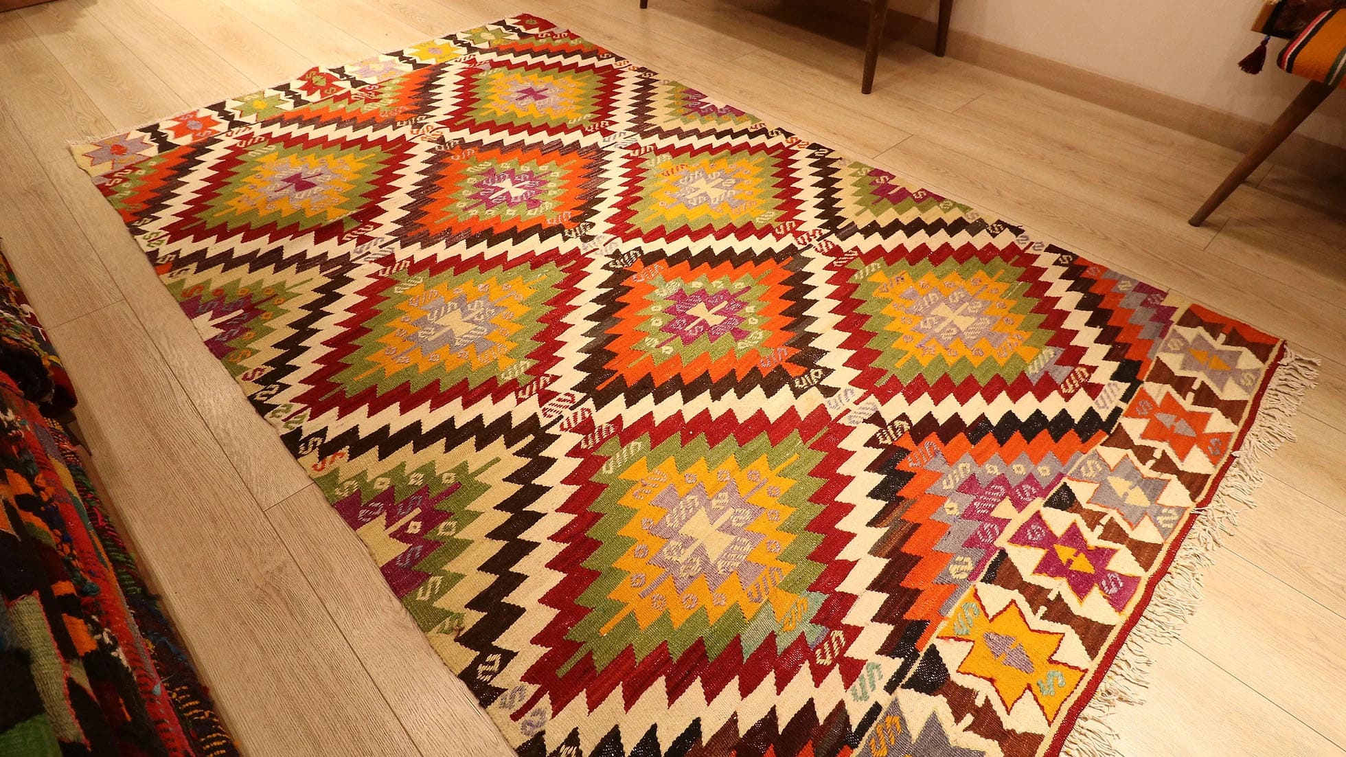 1960s handwoven kilim rug from Manisa, Turkey, 5x8 ft, featuring bold diamond motifs in pistachio, saffron, and clementine hues outlined in black on a white backdrop. The rug includes 'fetter' border motifs symbolizing connection and unity, with delicate tassels, blending cultural heritage with modern design.
