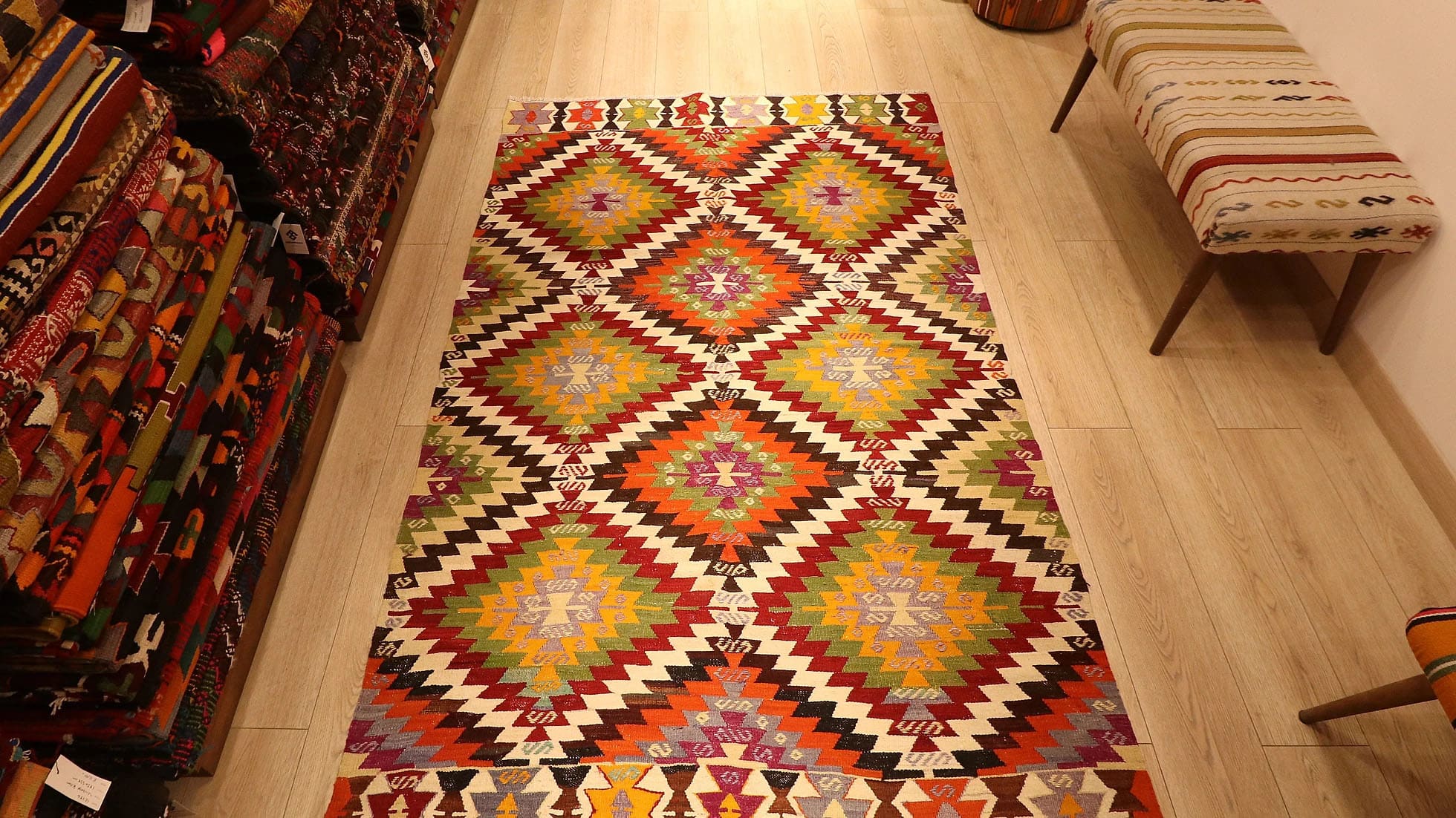 1960s handwoven kilim rug from Manisa, Turkey, 5x8 ft, featuring bold diamond motifs in pistachio, saffron, and clementine hues outlined in black on a white backdrop. The rug includes 'fetter' border motifs symbolizing connection and unity, with delicate tassels, blending cultural heritage with modern design.