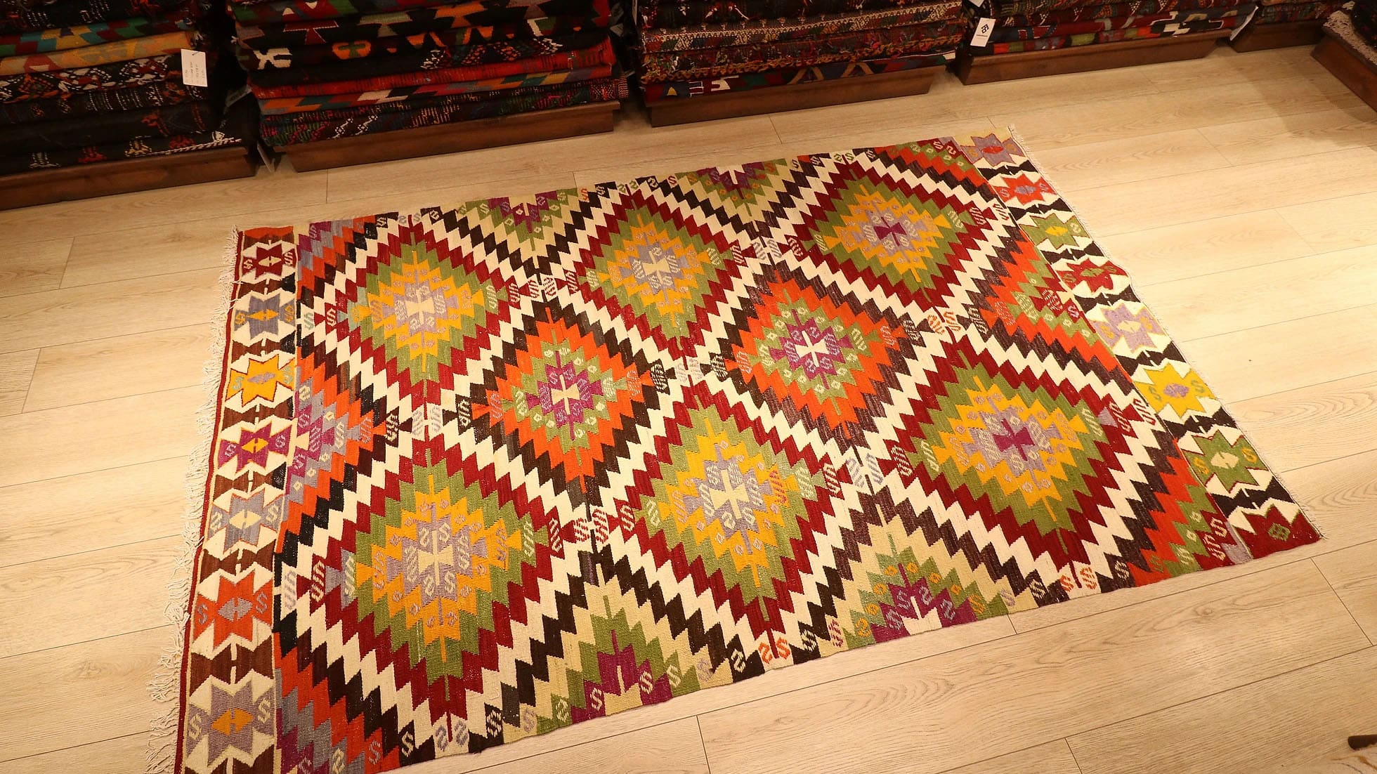 1960s handwoven kilim rug from Manisa, Turkey, 5x8 ft, featuring bold diamond motifs in pistachio, saffron, and clementine hues outlined in black on a white backdrop. The rug includes 'fetter' border motifs symbolizing connection and unity, with delicate tassels, blending cultural heritage with modern design.
