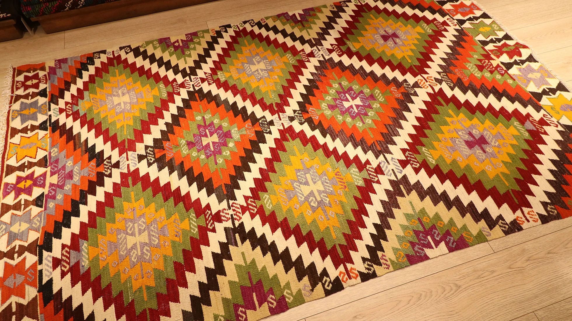 1960s handwoven kilim rug from Manisa, Turkey, 5x8 ft, featuring bold diamond motifs in pistachio, saffron, and clementine hues outlined in black on a white backdrop. The rug includes 'fetter' border motifs symbolizing connection and unity, with delicate tassels, blending cultural heritage with modern design.