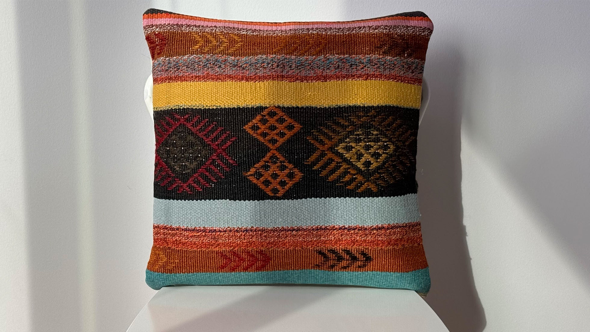 Vintage Turkish throw pillow with deep brownish-black center featuring burdock motifs, surrounded by earthy orange, yellow, Bordeaux, and pastel stripes in blue and terracotta. Handwoven wool kilim pillow by Kilim Couture New York, 16x16 inches, adds rustic elegance and cultural charm to any decor.