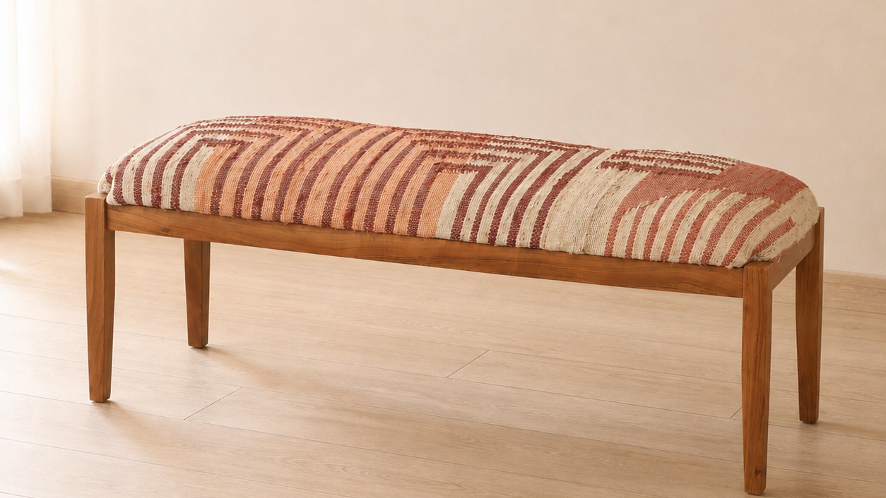 A wooden ottoman bench with a rectangular cushion upholstered in a rust, terracotta, and cream striped patterned kilim fabric, set against a neutral cream wall and light wood floor.
