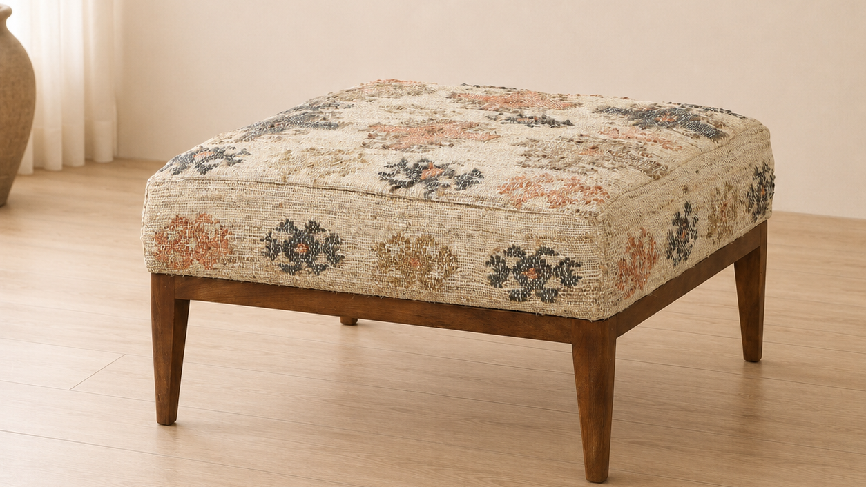 A square upholstered kilim ottoman with a kilim pattern in beige, rust, and navy, supported by a medium-brown wooden frame with tapered legs, set on a light wood floor.