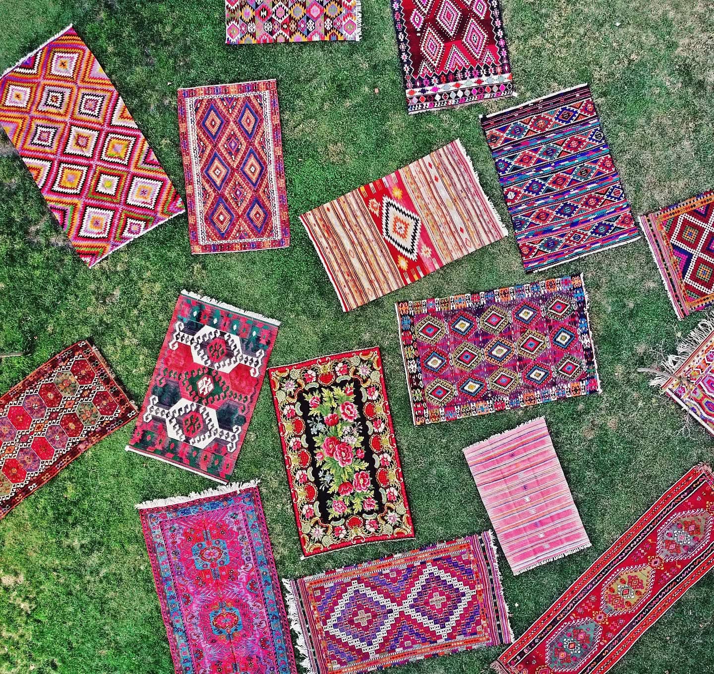 A collection of antique and vintage kilims laid out on lush green grass, showcasing vibrant geometric and floral patterns in shades of red, orange, pink, and blue.