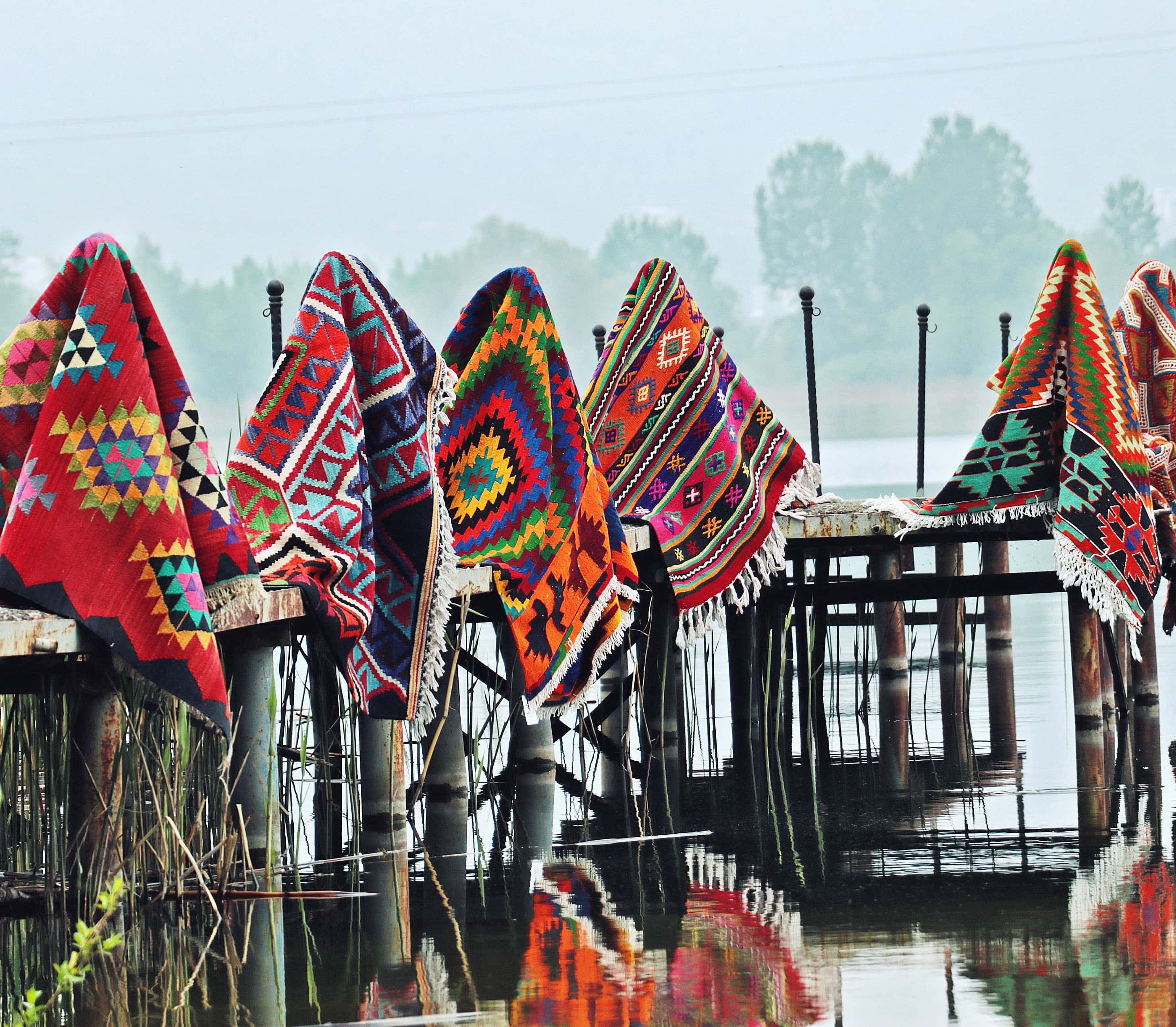 Handwoven kilims draped over a rustic wooden dock by a calm lake, reflecting their intricate tribal patterns in the water against a misty, serene background.