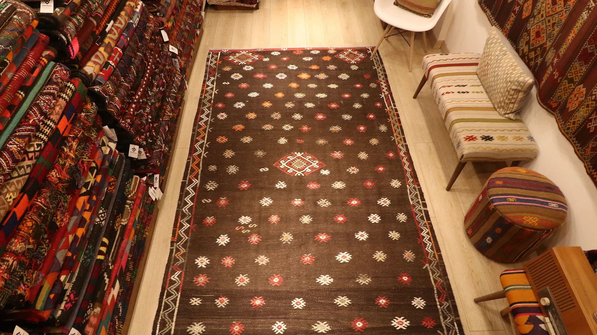 Vintage Turkish handwoven Balikesir tribal flat-weave rug from the 1950s in rustic taupe, featuring vibrant geometric patterns in red, orange, and white, displayed in a cozy showroom with stacked rugs, a kilim-upholstered bench, and matching kilim decor.